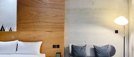 A modern hotel room with a wooden headboard, a bed with white linens, a bedside table with a lamp, and a bench with cushions.