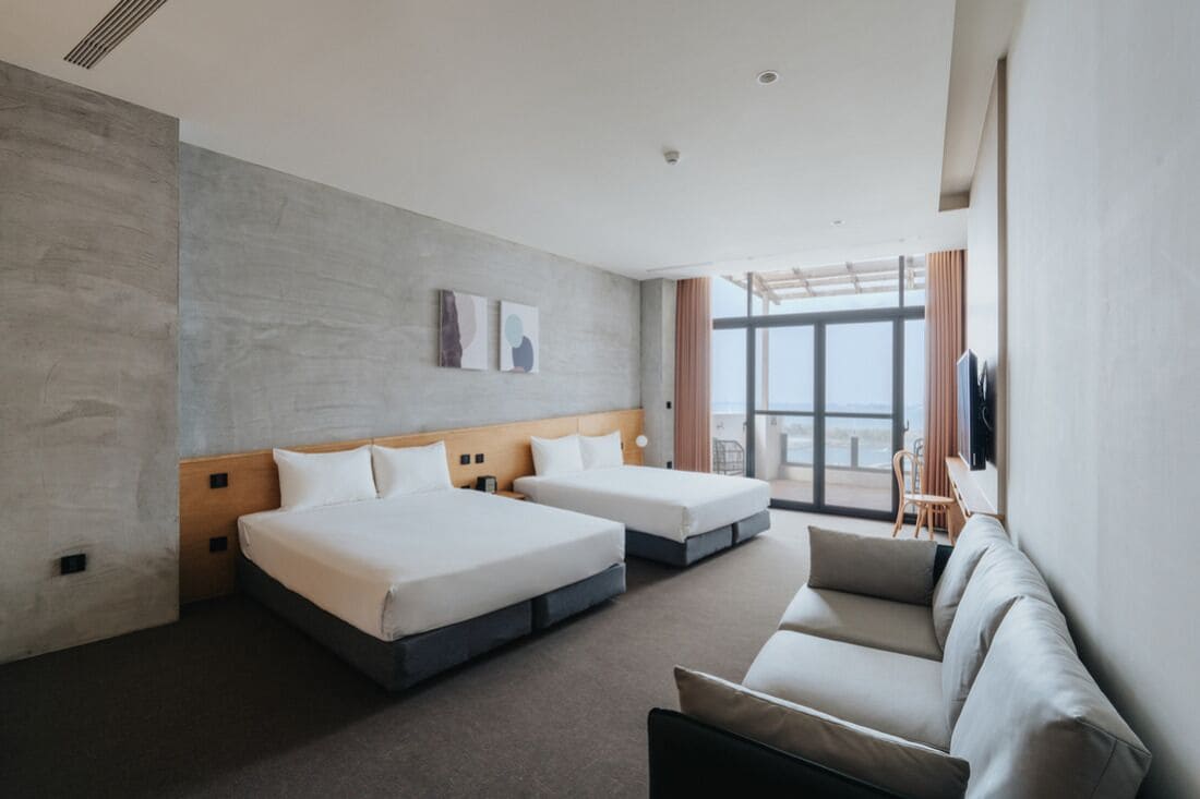 A hotel room with two beds, a sofa, and a large window offering a view.