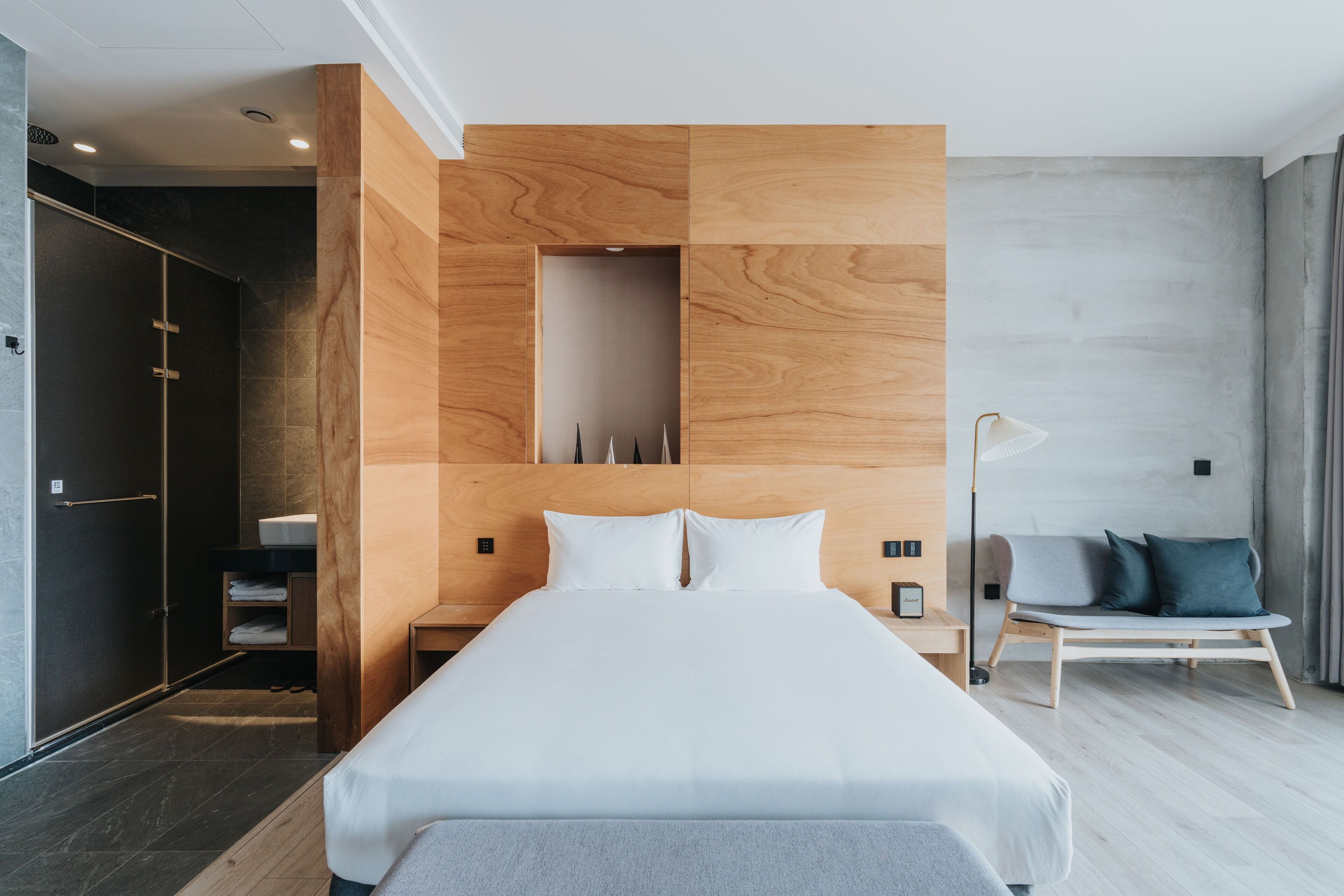 A modern hotel room with a large bed, wooden headboard, a bedside table, a nightstand, a chair, and a wardrobe.