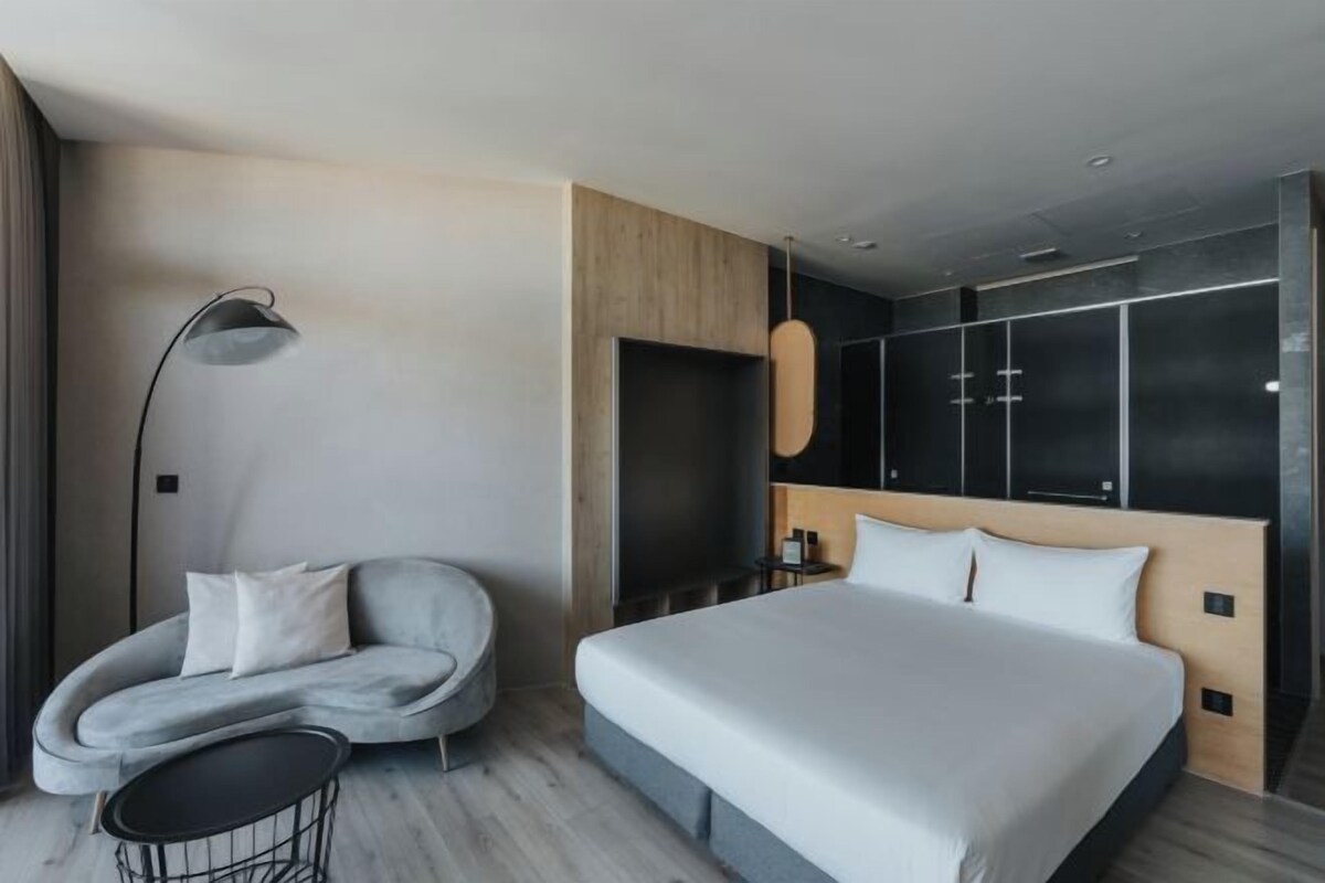 Una habitación de hotel moderna con una cama grande, un sofá gris curvo, una mesita redonda pequeña y una lámpara de pie.