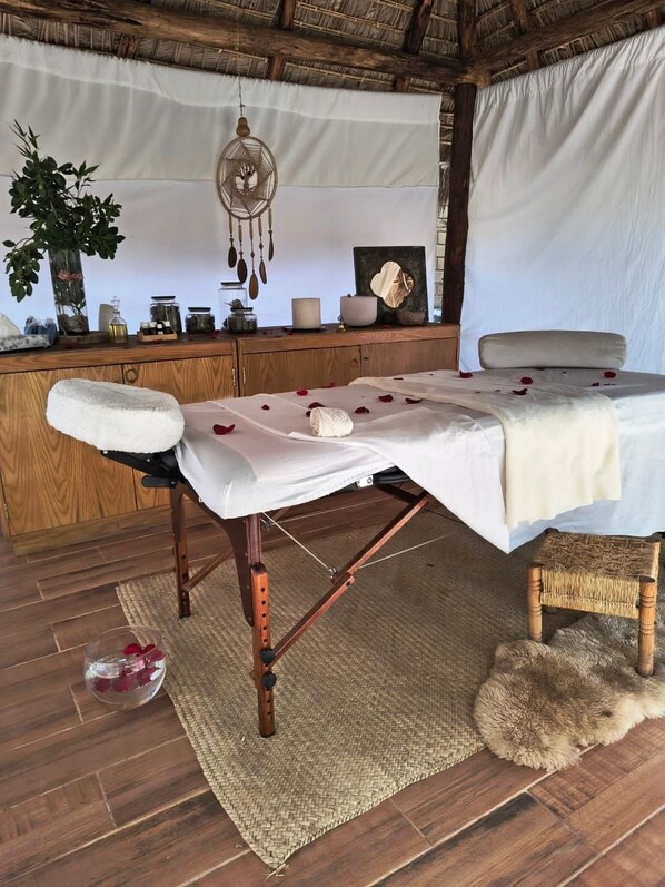 A massage table with a white sheet, a small wicker stool, a woven mat, and a dreamcatcher hanging above.