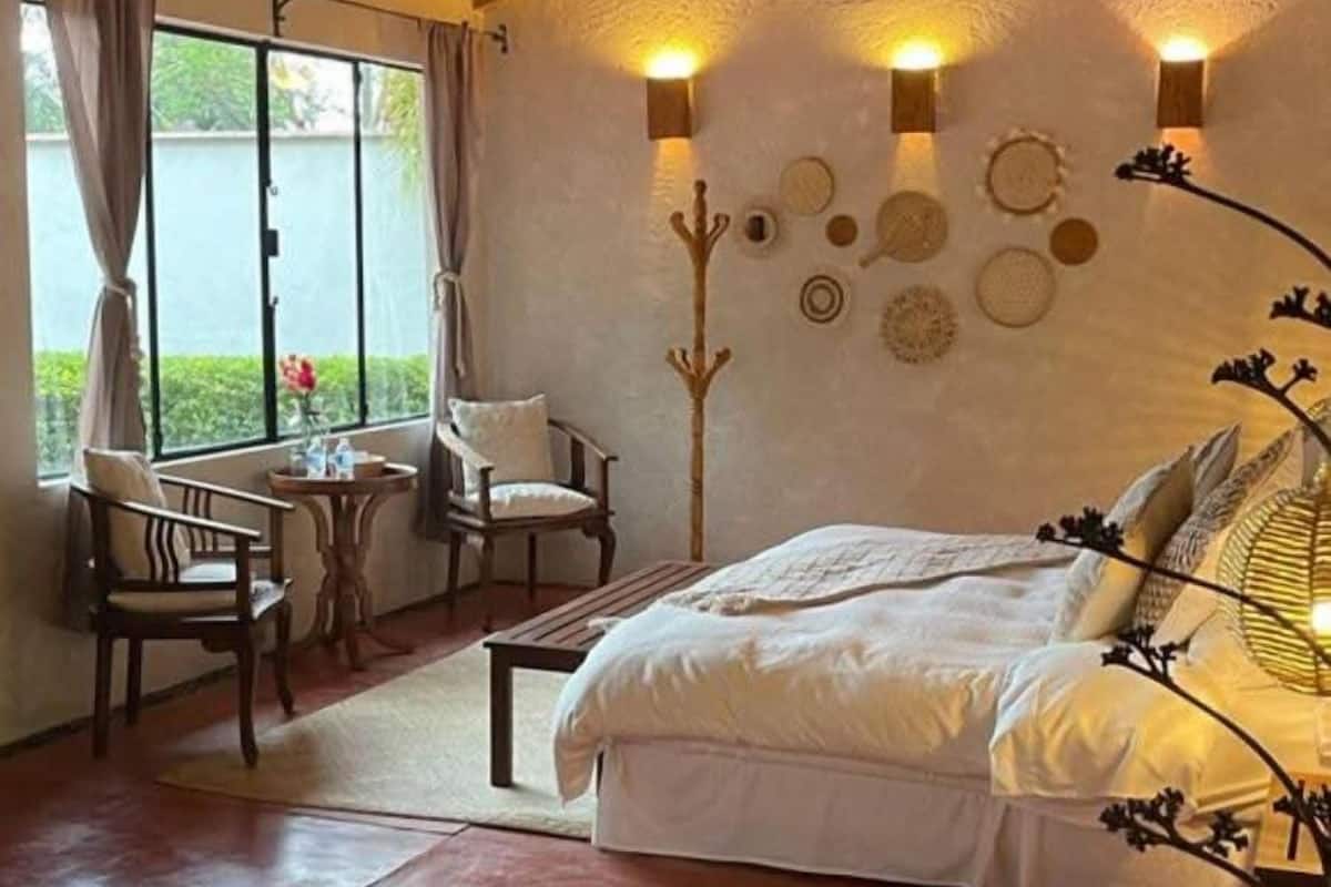 Quarto com cama, duas cadeiras, uma mesinha e um detalhe decorativo na parede.