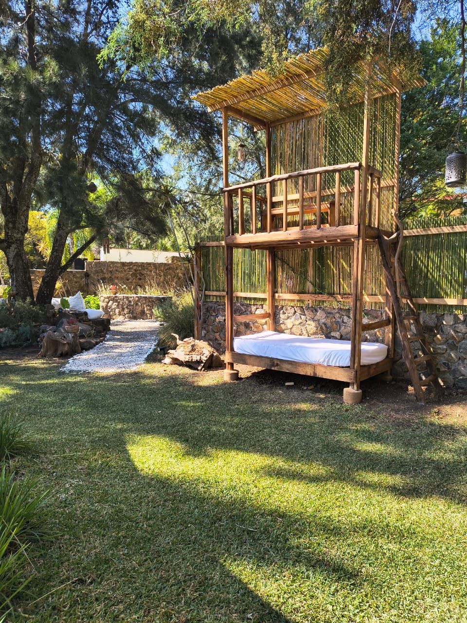Una casa sull'albero in legno con una piattaforma e un livello inferiore, circondata da un muro in pietra e vegetazione lussureggiante.