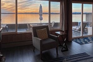 A room with a view of a body of water, a chair, a small table, and a window with curtains.