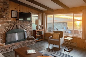A cozy living room with a brick fireplace, a television mounted above it, and a view of the sunset over the water.