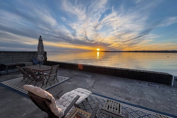 A sunset view from a terrace with a table and chairs, a covered umbrella, and a cushioned seating area.