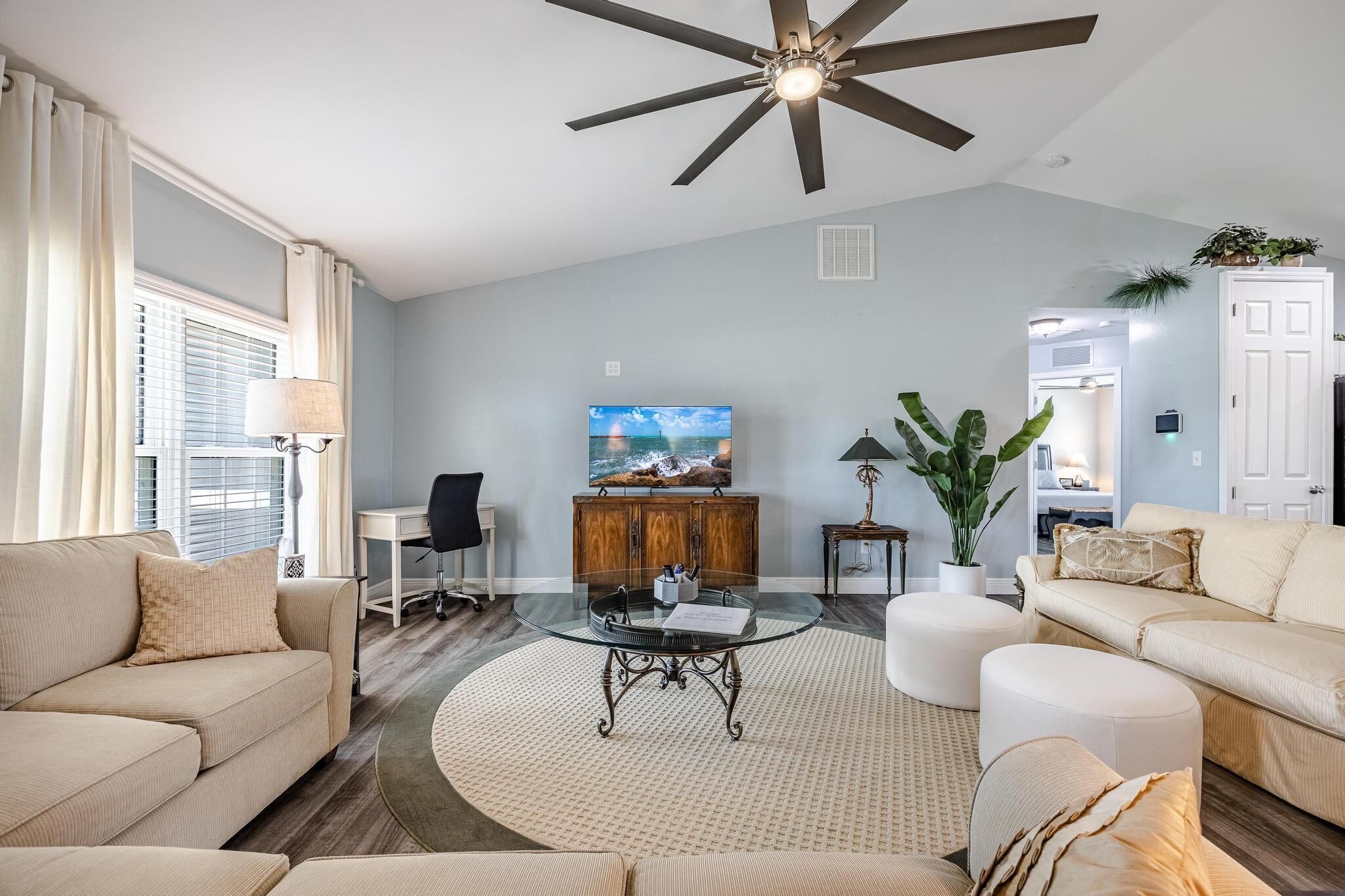 A living room with a glass coffee table, beige sofas, a ceiling fan, and a TV on the wall.