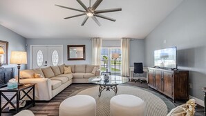 A living room with a sectional sofa, a glass coffee table, a ceiling fan, a TV, and a view of the outdoors through a window. - Perfect Getaway, Anytime of Year, Sunny, Clean, Spacious & Ideally Located! (Cape Coral)