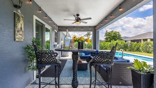 An outdoor patio with a table and two chairs, a ceiling fan, string lights, and a view of a swimming pool and greenery. - Perfect Getaway, Anytime of Year, Sunny, Clean, Spacious & Ideally Located! (Cape Coral)