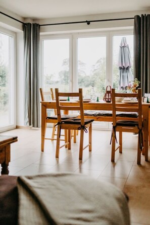 A dining area with a wooden table and chairs, large windows, and a view of the outdoors.