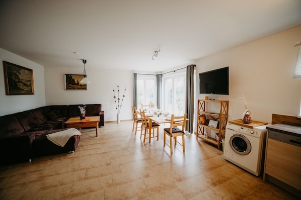 Ein Wohnzimmer mit einer Couch, einem Esstisch, Stühlen, einem Fernseher und einer Waschmaschine.