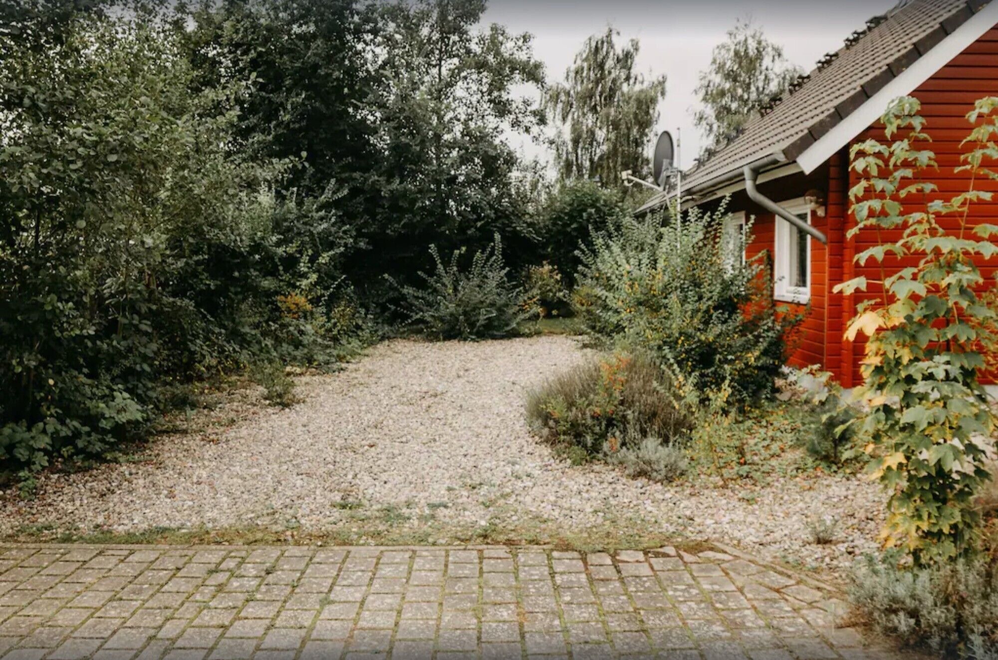 Un chemin de gravier mène à une maison rouge, entourée d’une végétation luxuriante.