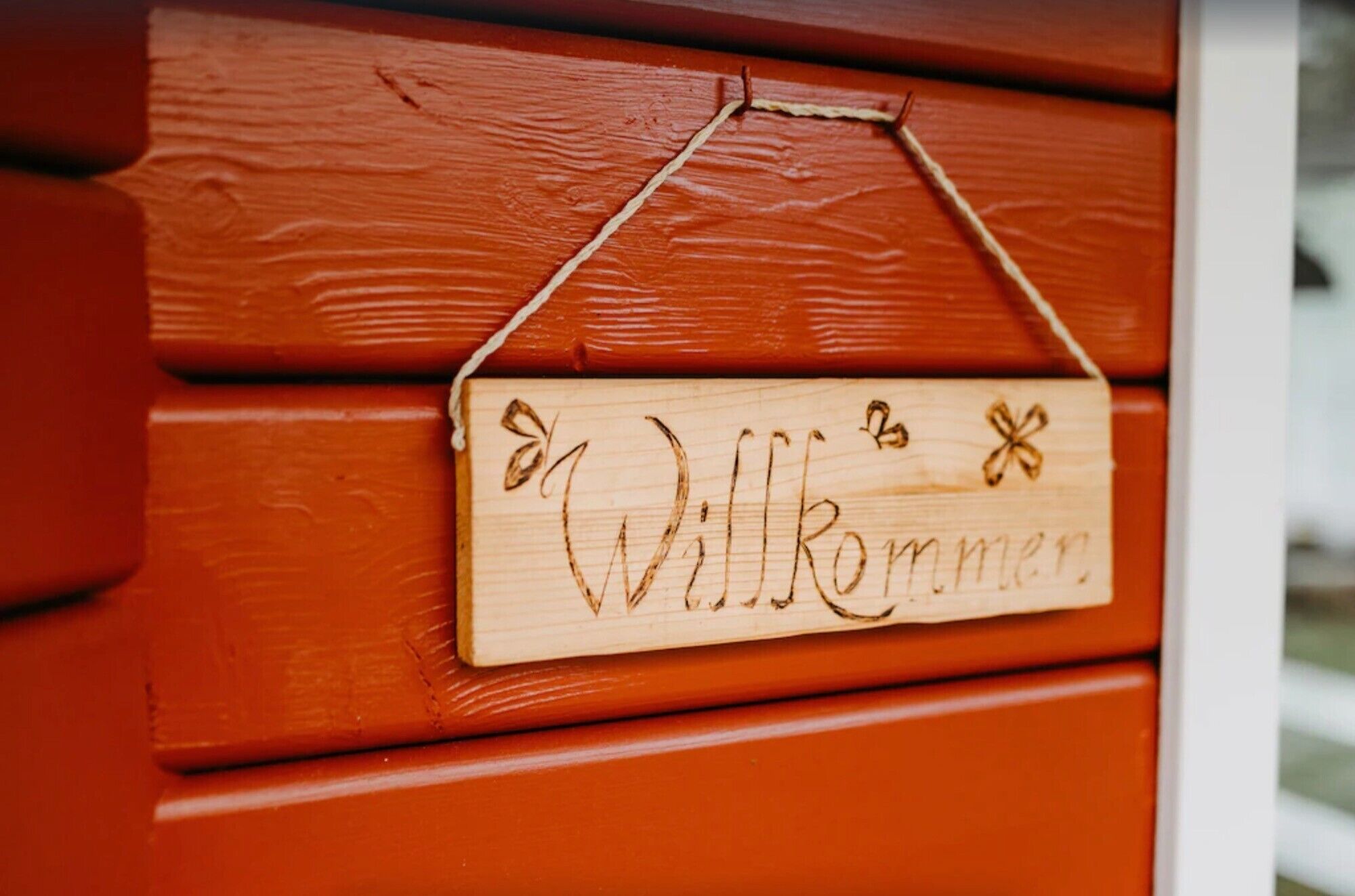 Ein hölzernes Schild mit dem Wort 'Willkommen' und dekorativen Elementen, an einer roten Holzfläche befestigt.