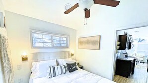 A bedroom with a bed, a ceiling fan, a window, a painting, and a door leading to a bathroom.