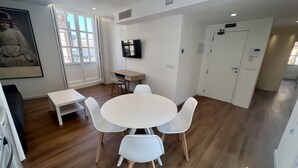 Un salon moderne avec une table ronde blanche et quatre chaises, un téléviseur à écran plat fixé au mur et une grande fenêtre donnant sur l’extérieur.