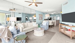 A modern living room with a sofa, coffee table, and a dining area with chairs. There is a kitchen area with a refrigerator and a microwave.
