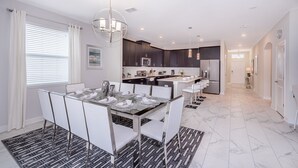 Una cocina moderna con armarios oscuros, una isla central y un comedor con mesa de vidrio y sillas blancas.