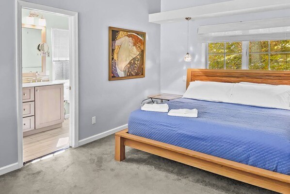 A bedroom with a bed, a bedside table, a window with a view of trees, and a bathroom with a sink and mirror.