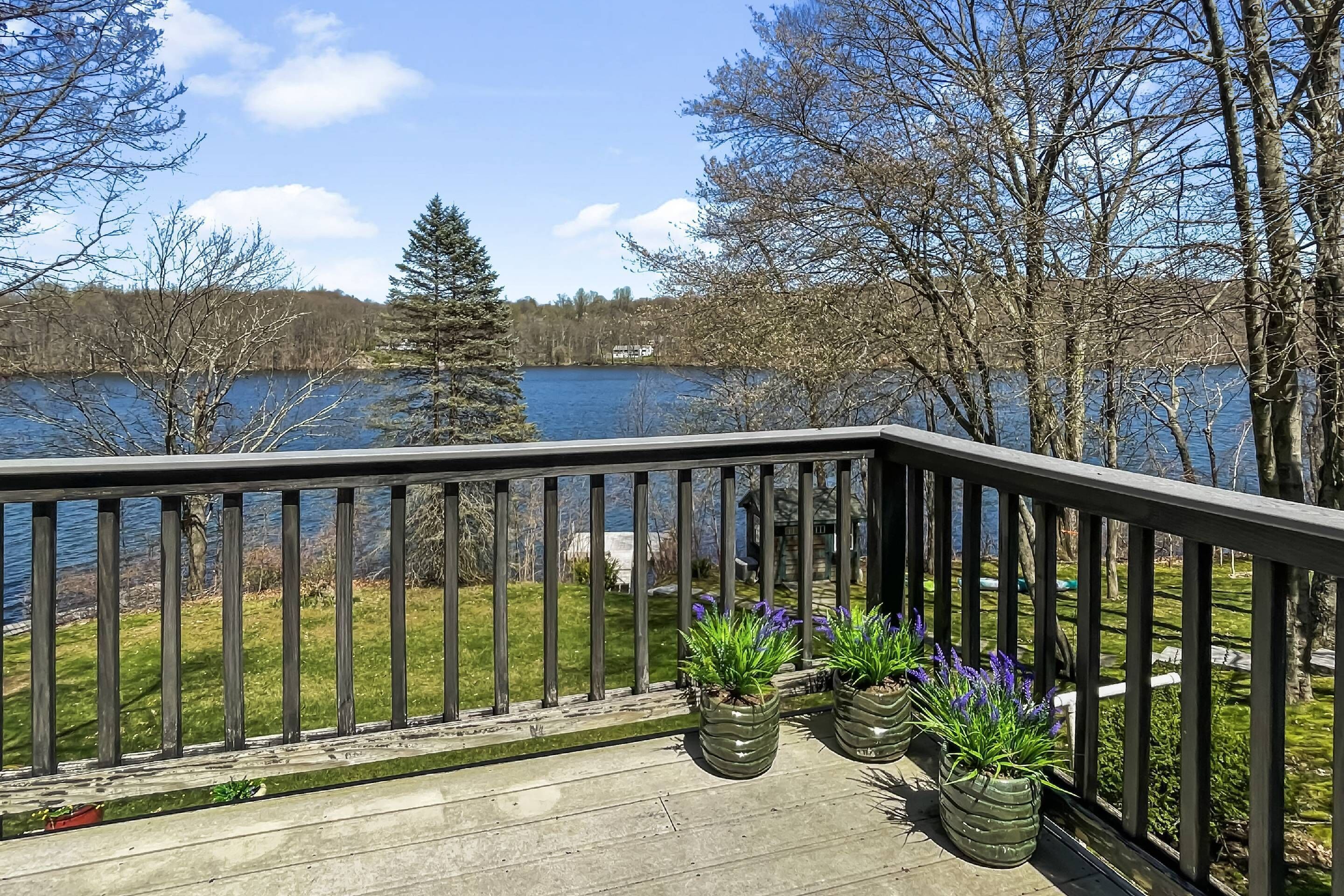 Um deck de madeira com guarda-corpo, de frente para um lago, cercado por árvores e plantas em vasos.
