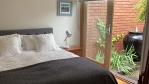 Ein Schlafzimmer mit einem Bett, einem Nachttisch, einer Lampe, einem gerahmten Bild und Blick auf eine Ziegelwand und Pflanzen durch eine Glastür.