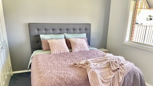 Une chambre à coucher avec une tête de lit rembourrée de couleur grise, une couvre-lit rose matelassée et plusieurs coussins.