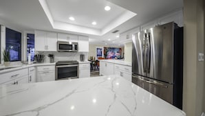 A modern kitchen with white cabinetry, a large marble island, stainless steel appliances, and recessed ceiling lighting. - Tropical 5BR Pool Beach House - 5min from Beaches! (Seminole)