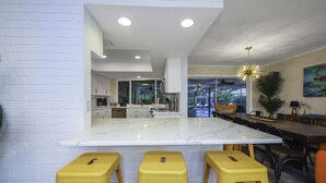 A modern kitchen with a marble island, yellow stools, and a dining area with a large table and chairs. - Tropical 5BR Pool Beach House - 5min from Beaches! (Seminole)
