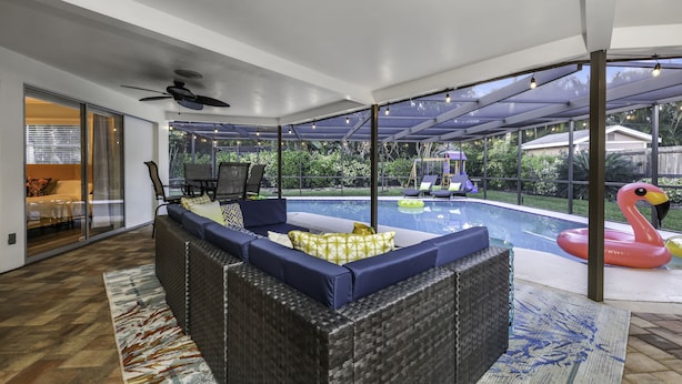 A modern living room with a large sectional sofa, a glass door leading to a pool area, and a ceiling fan. - Tropical 5BR Pool Beach House - 5min from Beaches! (Seminole)
