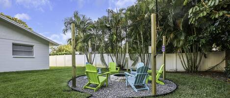 A backyard with a fire pit, green Adirondack chairs, and a white building.