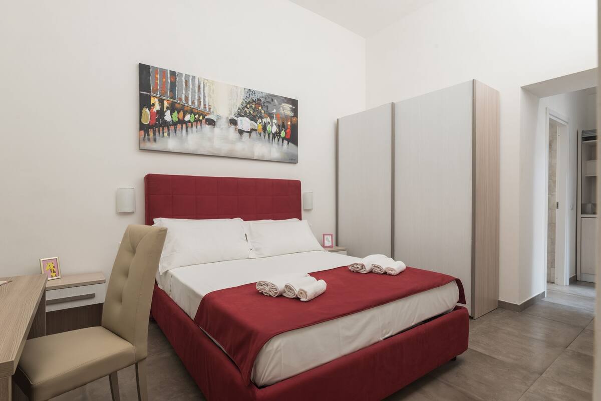 A hotel room with a bed, a chair, a desk, a painting on the wall, and a wardrobe.