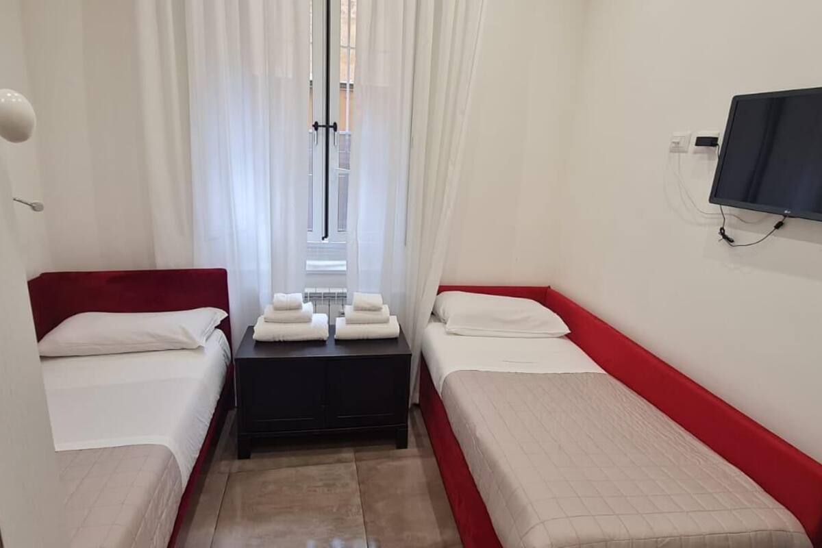 A hotel room with two single beds, a TV mounted on the wall, and a small table with towels.