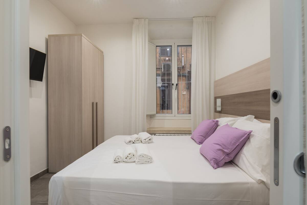 A hotel room with a bed, pillows, a wardrobe, and a window with curtains.