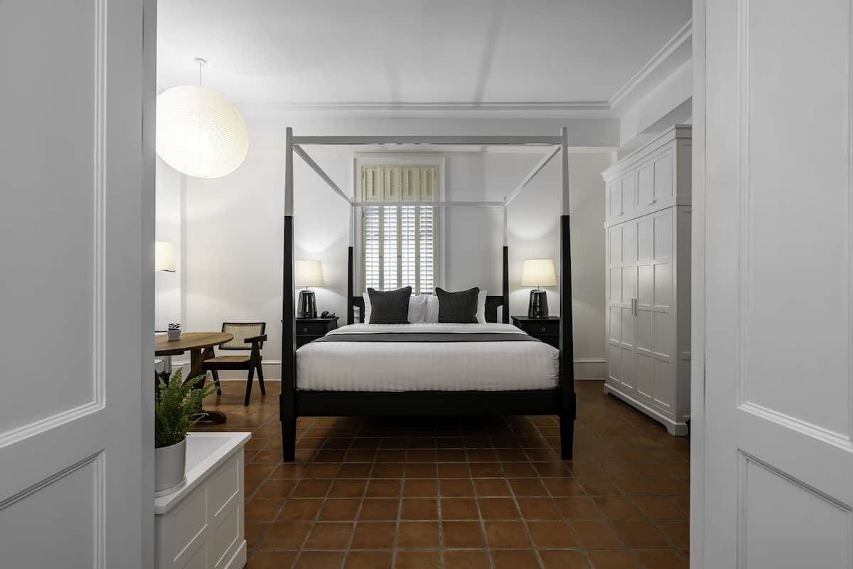 Une chambre avec un lit à baldaquin, deux tables de chevet avec des lampes, une fenêtre avec des stores et une plante en pot sur le côté gauche.