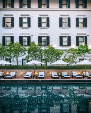 Een hotel met een zwembadgedeelte met ligbedden en parasols, een witte gevel en meerdere ramen met luiken.