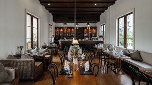 Un restaurant doté d’une longue table en bois, dressée pour le repas, d’un bar situé au fond de la salle et de grandes fenêtres laissant entrer la lumière naturelle.