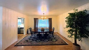 A dining area with a wooden table, chairs, and a chandelier. There is a window with curtains and a view of the outdoors. A potted plant is on the right side.