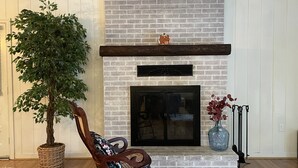 A living room with a brick fireplace, a wooden rocking chair, a potted plant, and a vase with flowers.