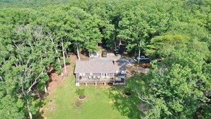 A two-story house surrounded by dense forest, with a well-maintained lawn and a driveway leading to a parked car.