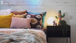 Un dormitorio moderno con pared de ladrillo blanco, una instalación de luces de neón, una cama con almohadas de colores, un edredón texturizado y un cactus en una mesita de noche.