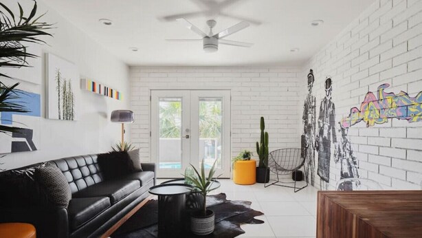 Una sala moderna con un sofá de cuero negro, una mesa de centro y una mesa de comedor. Hay plantas en macetas y un cactus en el suelo.
