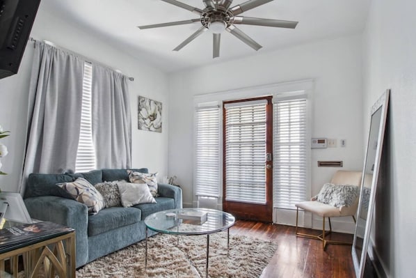 A living room with a blue sofa, a glass coffee table, a fluffy rug, a ceiling fan, and a door with blinds.