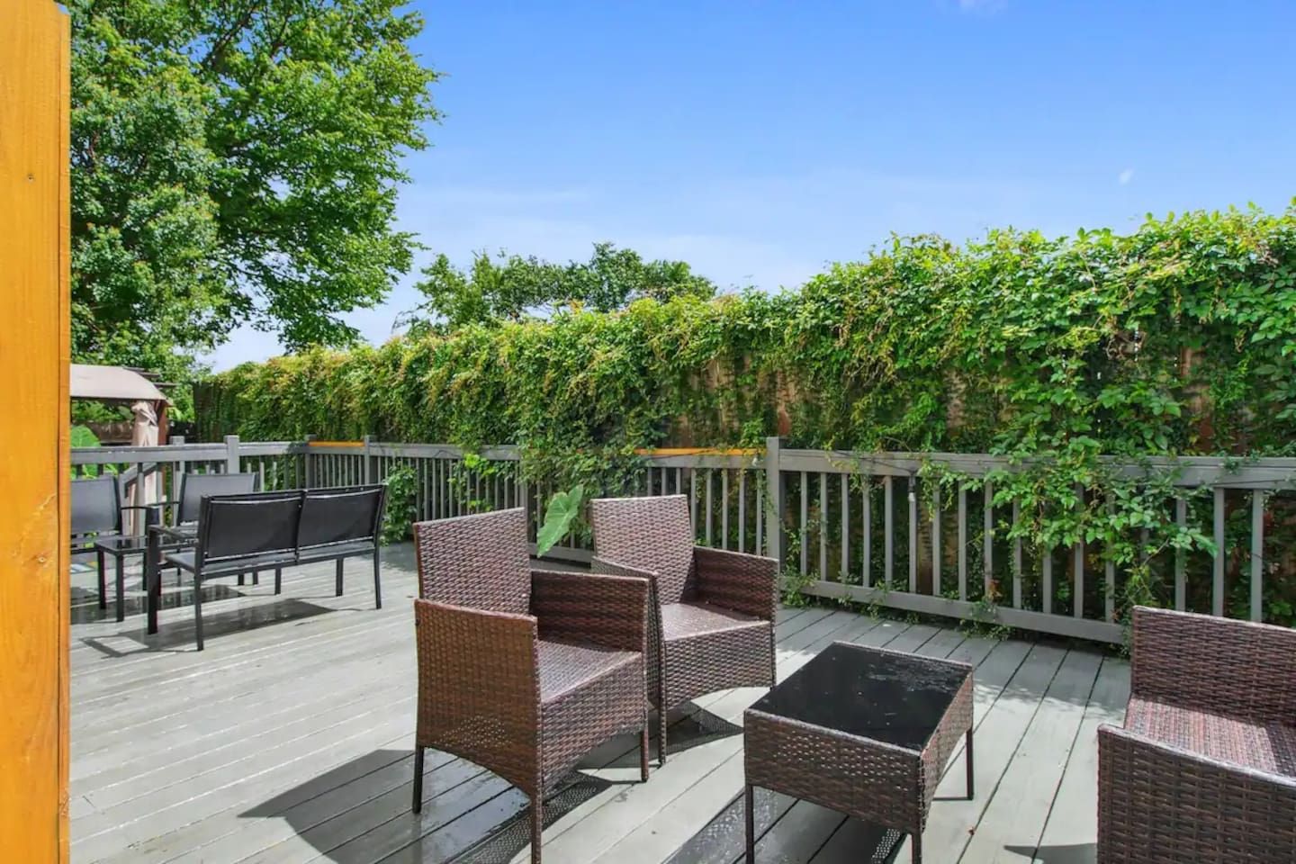 A wooden deck with wicker furniture and a glass table, surrounded by greenery.
