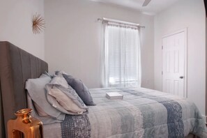 A bedroom with a bed, a bedside table, a lamp, a window with curtains, and a door.