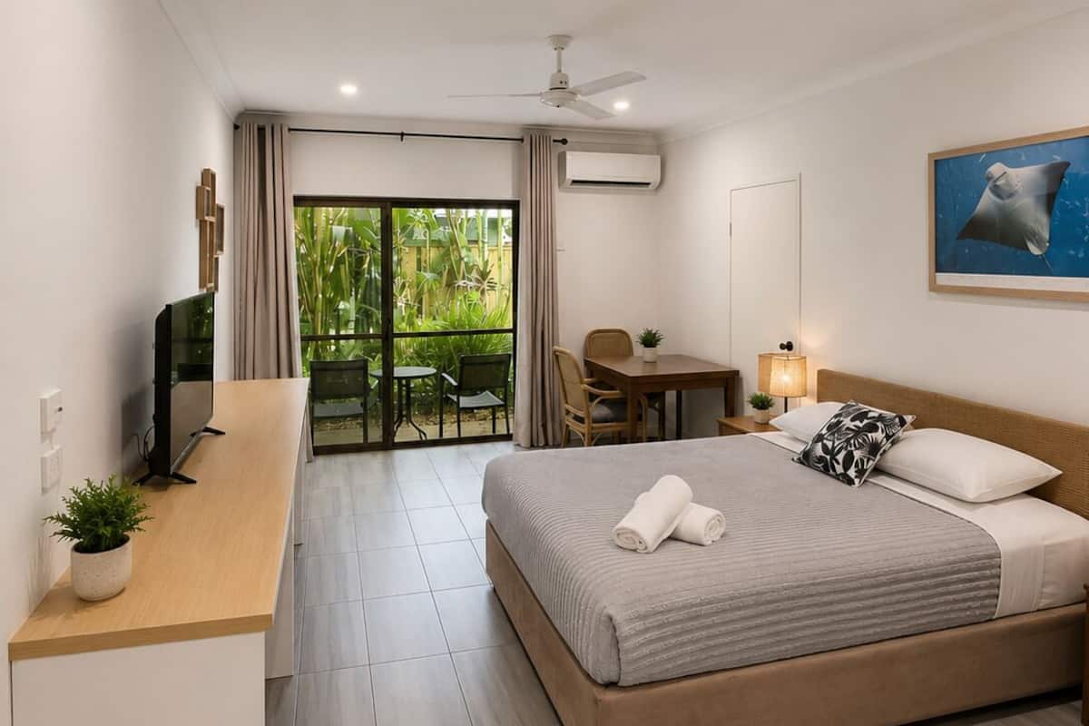 A hotel room with a bed, desk, chair, and a view of greenery.