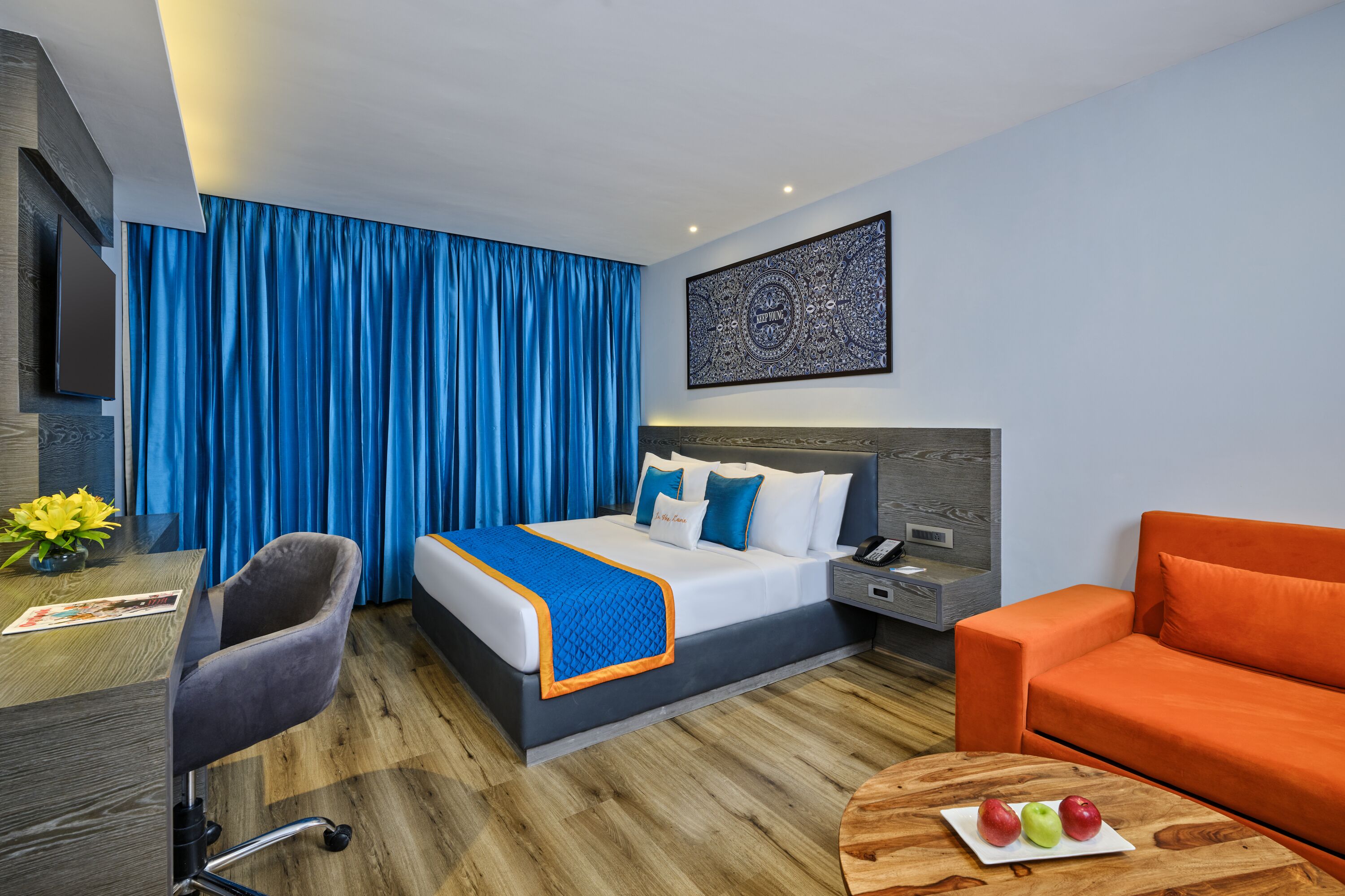 A modern hotel room with a bed, a desk, a chair, an orange sofa, and a coffee table with apples.
