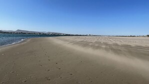 Ein weiter Sandstrand mit Fußabdrücken, die zum Wasser führen, ein klarer, blauer Himmel und Gebäude in der Ferne entlang der Küste.