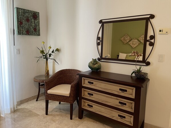 Una habitación con una silla de mimbre, un aparador de madera con cajones, un espejo y un florero con flores.