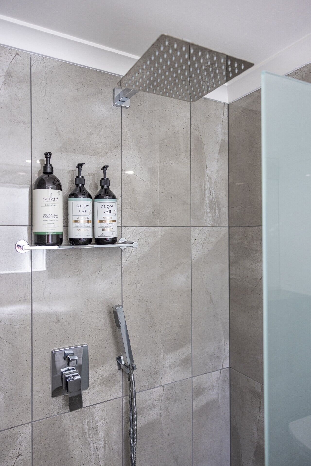 A modern bathroom with a shower, a glass door, and two bottles of SuKin body wash on a shelf.