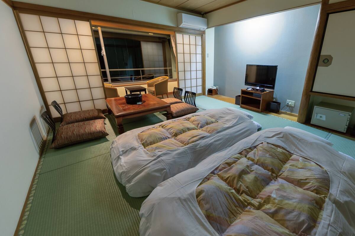 A traditional Japanese room with tatami flooring, two beds with futons, a television, a desk with a chair, and a sliding door.