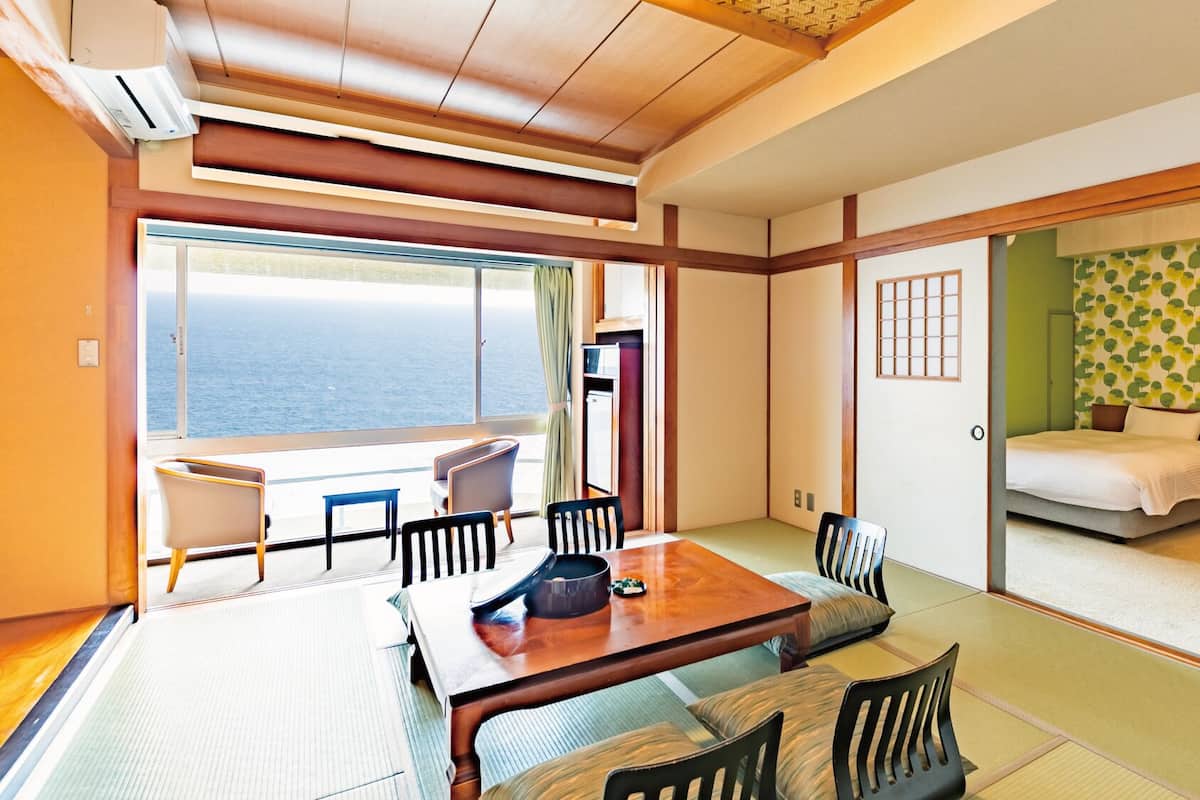 A room with a dining table, chairs, a bed, a wardrobe, and a view of the sea.